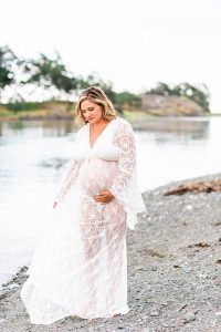 Pregnant woman walking on the beach holding her belly and flowing white maternity dress